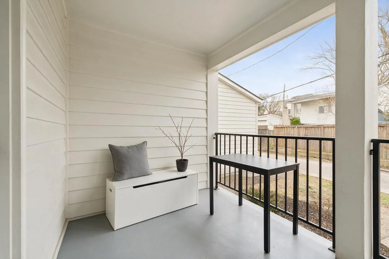 A small minimalist front porch with white horizontal siding, a light gray floor, and a simple black metal railing. A white storage bench with a gray cushion and small potted twig arrangement sits beside a black table, creating a clean and calm outdoor space.