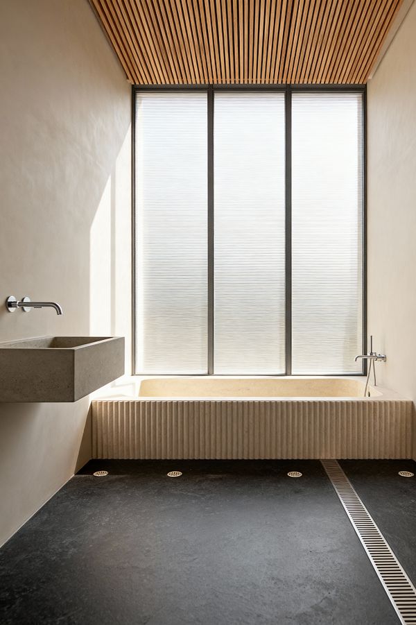 A minimalist bathroom featuring a wall of reeded translucent glass that diffuses daylight. A pale limestone soaking tub with a vertically fluted front sits beneath the window with a wall-mounted chrome filler, while a floating rectangular cast-concrete sink and spout occupy the left wall. The ceiling is lined with natural oak slats, and the floor is honed black stone with a stainless linear drain and round grates, framed by smooth warm off-white plaster walls.