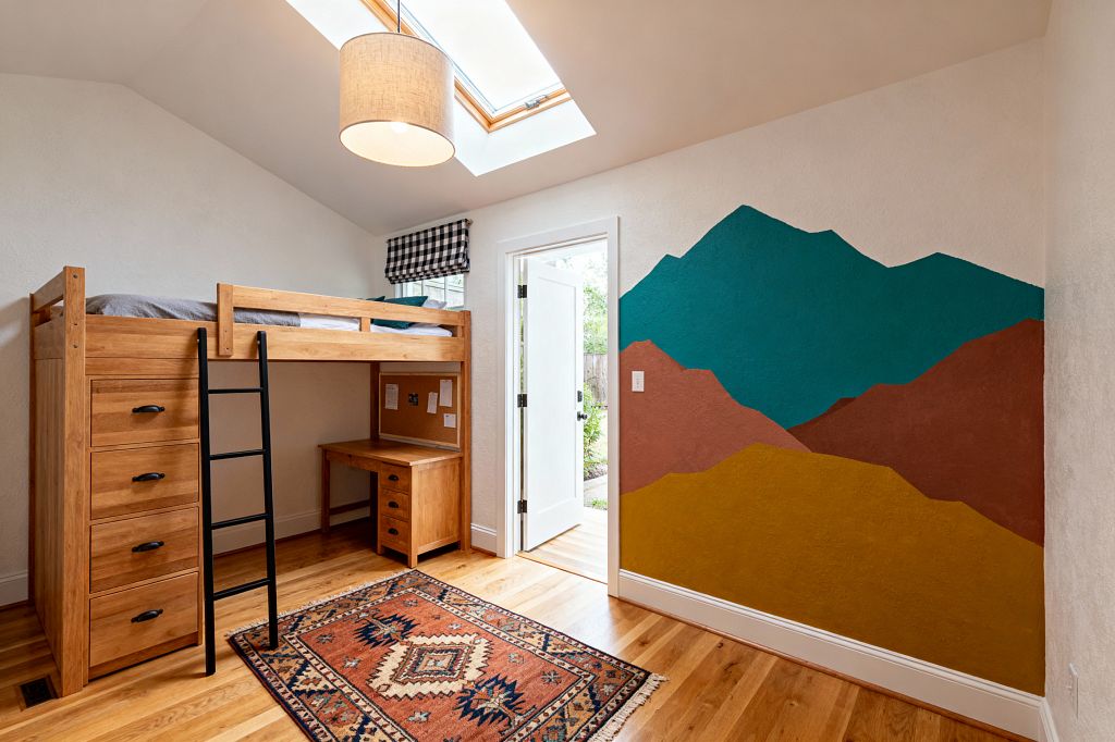 Kids’ room with a lofted oak bed featuring four drawers, a built-in desk, and a matte black ladder, set against warm white textured walls. A large skylight and a linen drum pendant illuminate a colorful geometric mountain mural in teal, terracotta, and mustard; a black-and-white gingham roman shade tops a small window, while white oak floors and a rust, navy, and cream patterned rug add warmth near a door opening to a garden.