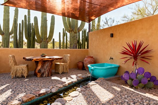 A sunlit Southwestern patio features an adobe stucco wall, a rust-colored shade sail, and desert cacti beyond. A live-edge wood table with two raffia-fringed chairs sits on pebble aggregate paving beside a turquoise basin under a wall spout that feeds a stone-lined rill; terracotta pots, a burgundy spiky plant, and a purple prickly pear add saturated color.