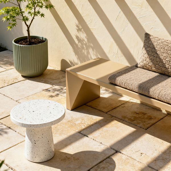 A modern patio vignette shows a sand-beige metal bench with a geometric leg and a chunky taupe woven cushion. A round white terrazzo pedestal side table sits on warm travertine tiles, while a ribbed sage-green planter holds a small tree against a textured cream stucco wall. Strong sunlight creates diagonal shadows across the wall and seating, emphasizing clean lines and a calm, minimalist look.