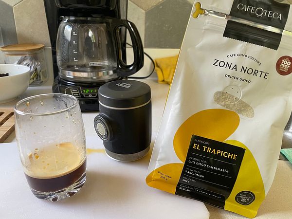 A close-up of a Wacaco espresso maker pouring a small cup of dark espresso on a wooden table with coffee beans and a bag of coffee.