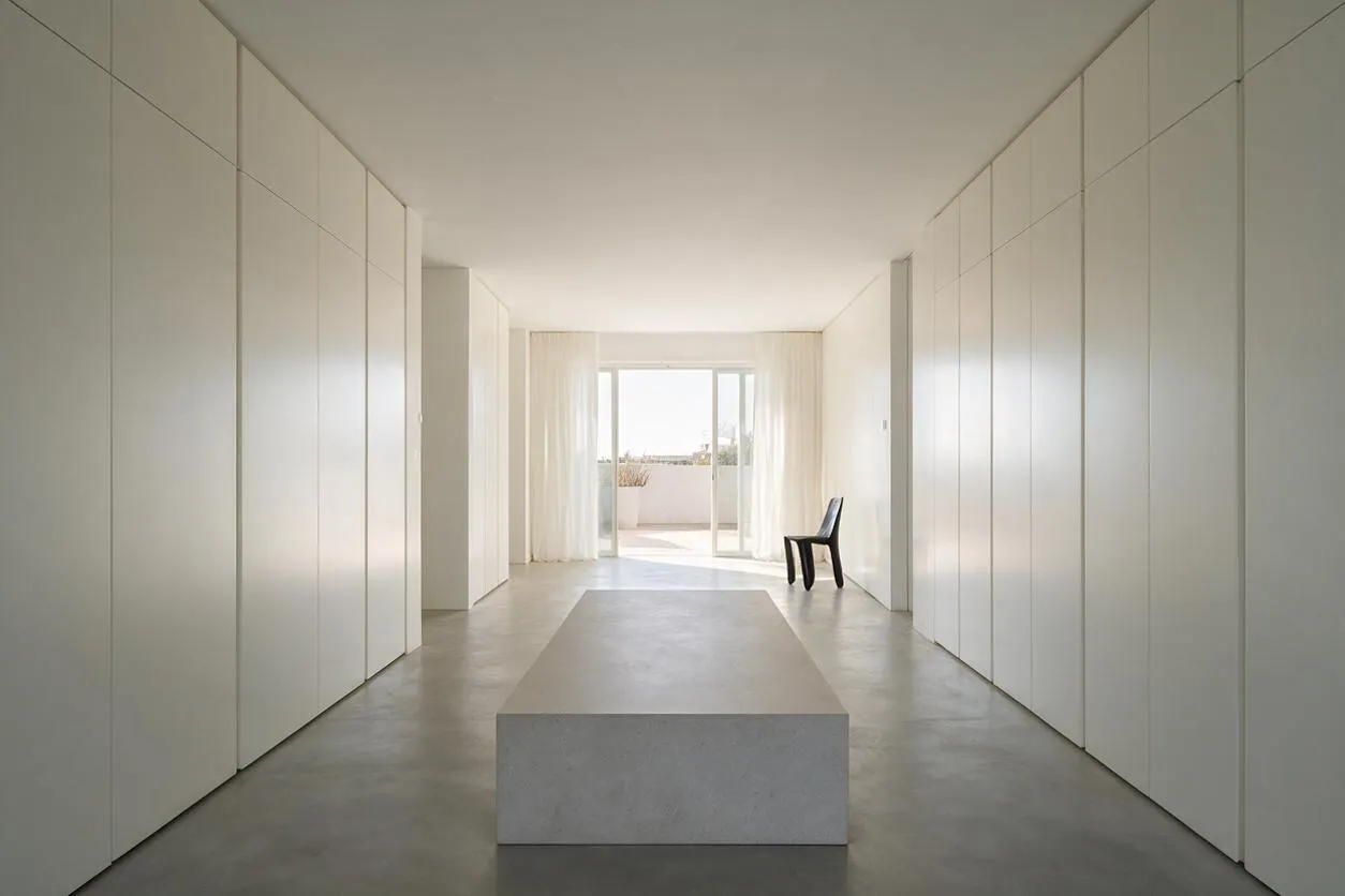 A long minimalist closet with floor-to-ceiling white cabinet doors lining both sides of a narrow room. A simple rectangular bench sits in the center, with a black chair near bright sliding doors and sheer curtains at the far end.
