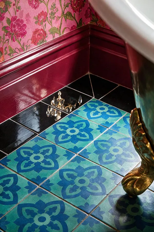 A maximalist bathroom corner with turquoise patterned floor tiles, glossy black border tiles, deep burgundy trim, and pink floral wallpaper above the wainscoting. A curved gilded furniture leg and crystal hardware add ornate detail and a richly layered, decorative feel.
