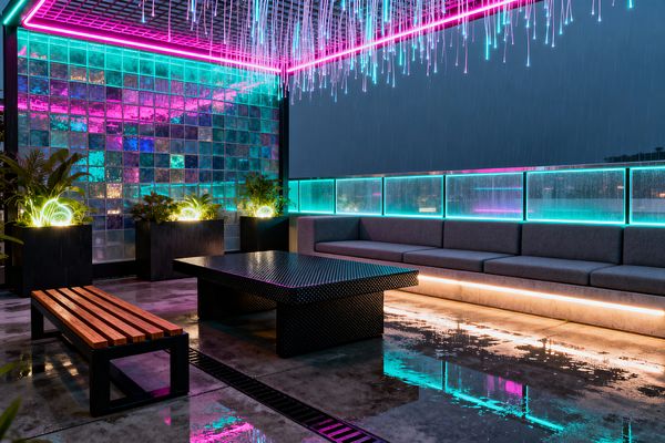 Nighttime rooftop patio in the rain, with polished concrete reflecting neon cyan and magenta lights. A ceiling grid drips fiber-optic strands, a long gray upholstered sectional sits on a concrete base with underlighting, and a perforated black metal coffee table faces a nearby wooden-slat bench. At left, an iridescent glass-block wall and matte black planters with glowing ring lights frame the space, while backlit glass railing panels edge the perimeter.