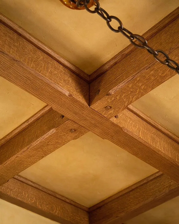A close-up view of a Craftsman-style ceiling showing intersecting exposed wooden beams with visible joinery and small decorative pegs. The beams frame warm beige plaster panels, and a dark metal chain from a ceiling fixture crosses the upper corner.