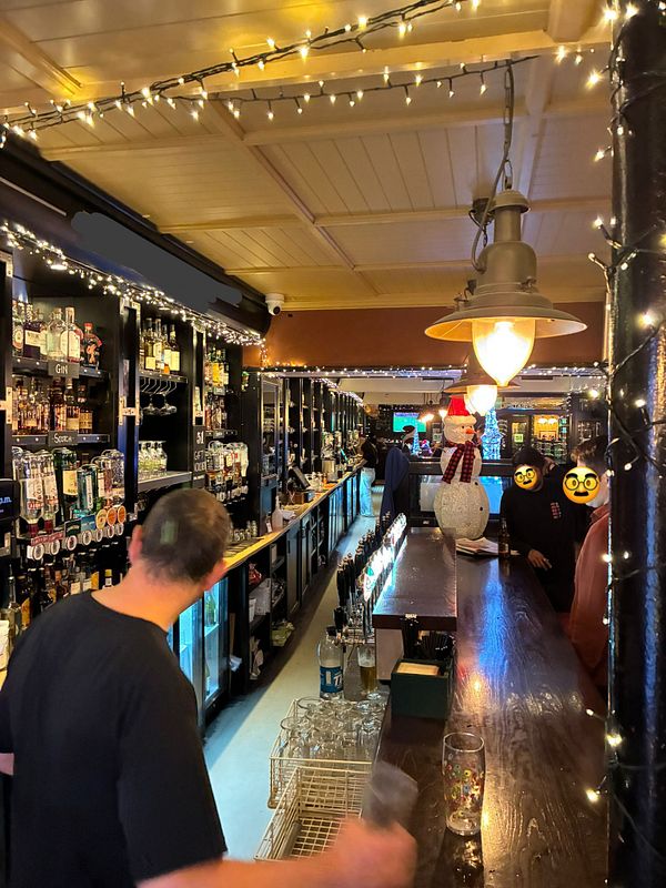 A lively pub interior with a bar, shelves of drinks, and festive decorations.
