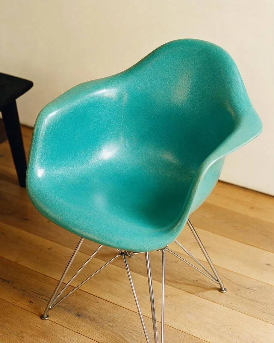 A turquoise molded plastic accent chair with an organic shell shape sits on thin chrome wire legs over a light wood floor. The curved seat has a glossy finish, and a plain cream wall and a partial black table appear in the background.