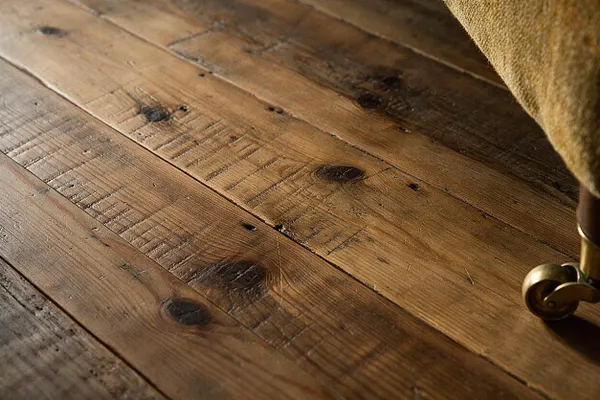 Close-up of wide rustic wooden floor planks with visible knots, grain, scratches, and aged texture in warm brown tones. A small brass wheel and upholstered furniture edge appear at the right side, reinforcing the cozy, lived-in setting.