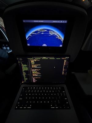 A laptop displaying code is positioned in front of an in-flight entertainment screen showing a map of the flight route to Tokyo.