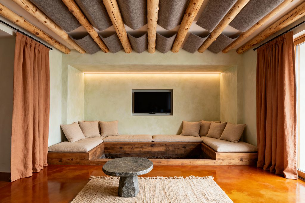 A Scandinavian-style media room features a recessed TV within a limewashed niche above a U-shaped built-in sofa with pine bases and beige linen cushions. Exposed round timber beams are spanned by gray felt acoustic panels; rust-colored linen curtains frame a large window, a rough stone round coffee table sits on a textured rug, and the warm terracotta floor glows under soft concealed LED lighting.