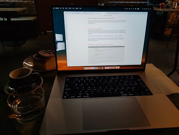 A laptop is open on a table with a cinnamon scroll, coffee, and water in a cafe setting.