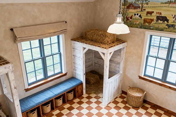 Bird’s-eye view of a farmhouse-themed kids’ room featuring a small barn play stall built from distressed whitewashed boards, with straw inside and a hay bale on top. Two slate-blue windows with white trim sit above a long wooden bench with a denim-blue cushion and cubbies holding woven baskets. The walls have a tan plaster finish, the floor is a terracotta-and-cream checkerboard, and a white enamel pendant hangs near a pastoral mural of cows and barns.
