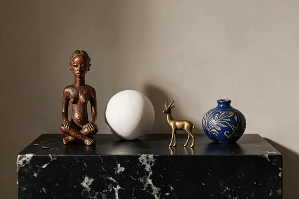 A curated tabletop vignette sits on a black marble pedestal against a warm beige wall. The display includes a carved wooden seated figure, a matte white sphere, a small brass deer, and a cobalt blue vase with gold botanical detailing.