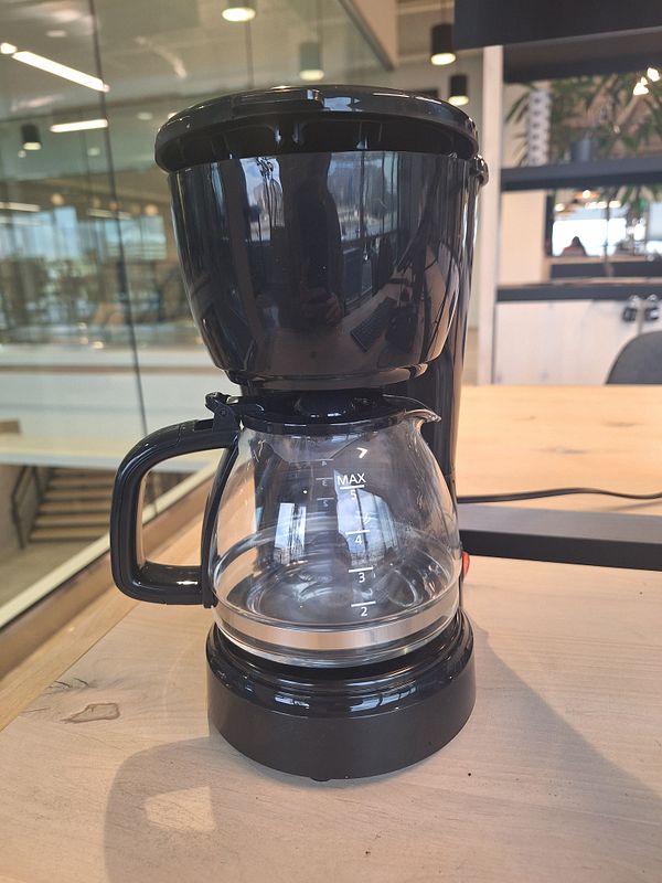 A compact black coffeemaker is displayed on a wooden table in a modern workspace.