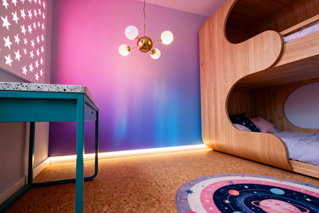 A kids’ bedroom in a space theme features a pink-to-indigo gradient wall illuminated by LED strip lighting at the base. A light oak built-in bunk bed with soft curved openings and a circular porthole holds starry and pastel bedding. A brass sputnik ceiling light with opal globes hangs above. A teal metal desk with a terrazzo top sits near a backlit star-cut panel, and a round galaxy rug rests on cork flooring.
