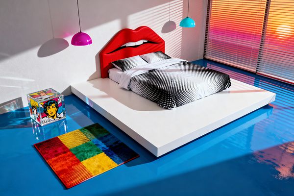 A pop art bedroom featuring a low white platform bed with a bright red, lip-shaped headboard and black-and-white halftone bedding. The glossy electric-blue floor reflects a magenta and a turquoise dome pendant. A clear acrylic cube with comic-style portraits serves as a nightstand beside a small rainbow patchwork rug, while horizontal blinds cast pink and orange light bands across the white wall and floor.