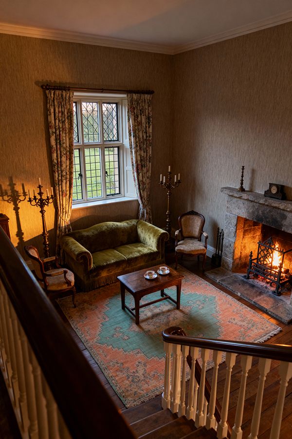 A traditional living room viewed from a staircase shows a stone fireplace with a bright fire, a wrought-iron grate, and fireplace tools, opposite a classic seating group. An olive green velvet sofa beneath leaded windows with floral curtains pairs with two carved armchairs, tall metal candelabras, and a small walnut coffee table set with teacups on a faded peach-and-seafoam rug over dark hardwood floors; warm beige textured walls and a mantel clock complete the scene.