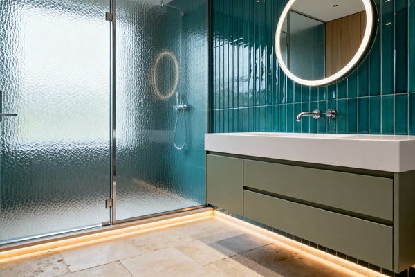 A contemporary bathroom features vertically stacked teal glazed wall tiles, a floating olive vanity with a white integrated basin, and a circular backlit mirror. A walk-in shower is enclosed by a hammered translucent glass screen with slim chrome hardware and a wall-mounted shower set; warm LED strips under the vanity illuminate a light beige travertine floor, creating a calm, spa-like atmosphere.