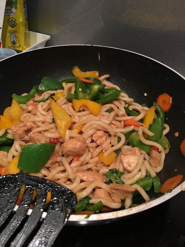 A stir-fried udon dish with colorful vegetables and chicken pieces in a frying pan.