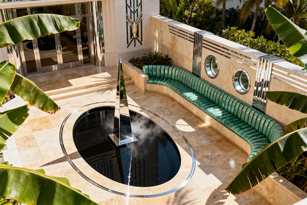 A sunlit Art Deco patio features warm travertine flooring and walls surrounding an oval reflecting pool edged in metal, centered by a mirrored stainless-steel obelisk emitting a light mist. A curved, ribbed emerald-green bench lines one side above a stone base, opposite chrome-rimmed porthole windows and horizontal metallic banding. Geometric door details and lush banana leaves frame the scene, creating a glamorous, streamlined outdoor lounge.