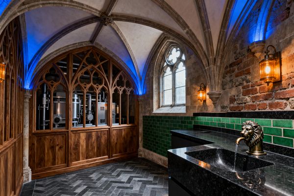 A Gothic-style laundry room features a ribbed stone vaulted ceiling with blue uplighting and warm lantern sconces. A carved wood tracery screen reveals stainless laundry equipment, while black granite counters with an undermount sink and a brass lion-head faucet sit before emerald green glazed tiles and an exposed brick wall; a leaded arched window and dark herringbone slate floor complete the dramatic, historic-meets-industrial space.