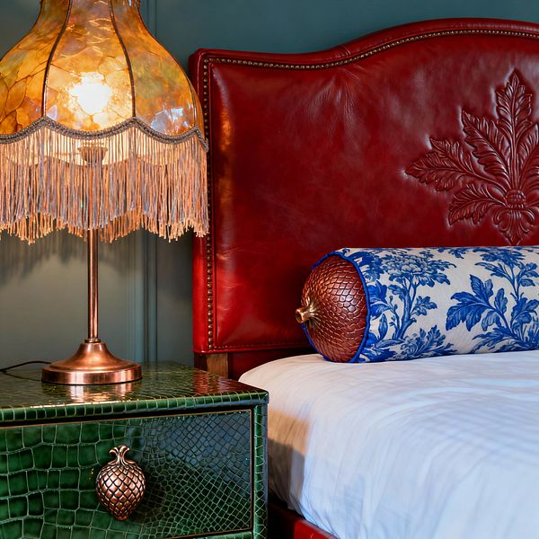 A richly layered bedroom vignette shows a warm-glowing fringed lamp on a green crocodile-textured nightstand with a copper pineapple pull beside a deep red leather headboard embossed with a large leaf and trimmed with nailheads. A white duvet is paired with a blue floral bolster featuring copper snakeskin end caps, all set against moody teal walls for dramatic contrast and tactile abundance.