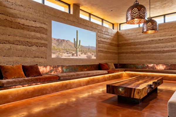 A Southwestern-style media room with rammed-earth walls and narrow clerestory windows shows a desert landscape projected directly onto the wall. A continuous built-in bench wraps the room with a patinated copper backrest, suede cushions and pillows in rust, terracotta, and sand, underlit by warm LEDs; a polished terracotta concrete floor and a live-edge hardwood coffee table anchor the space, while two perforated copper pendants cast patterned light for an intimate, earthy atmosphere.