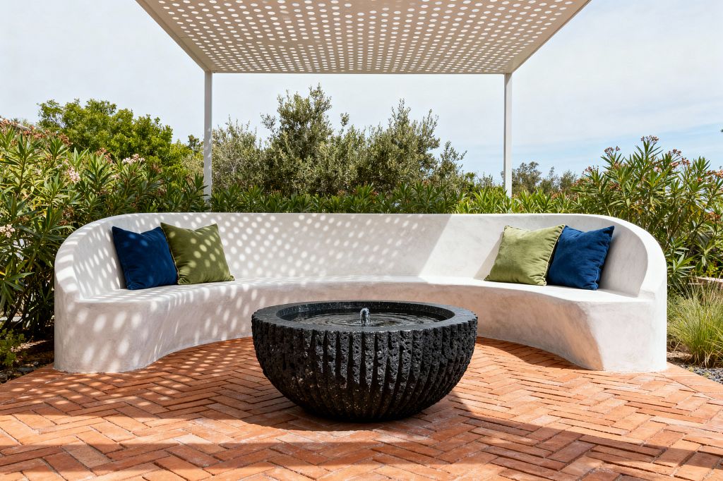 A modern outdoor patio features a smooth white, curved built-in stucco bench arranged in a semicircle beneath a perforated white metal canopy that casts circular shadows across the seating. At the center sits a low black textured stone bowl fountain with a small bubbling spout on warm herringbone terracotta paving, surrounded by dense shrubs and trees, with olive green and deep navy pillows adding color to the minimalist setting.