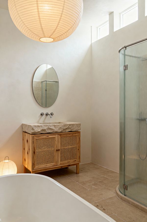 Light-filled Scandinavian bathroom with off-white plaster walls and high clerestory windows. A knotty pine vanity with woven cane doors supports a raw-edged limestone sink beneath a round mirror and wall-mounted stainless faucet. A curved corner shower features reeded glass and metal hardware, beige stone-look tiles cover the floor, a freestanding tub edge is visible in the foreground, and paper lantern lighting provides warm, diffused illumination.