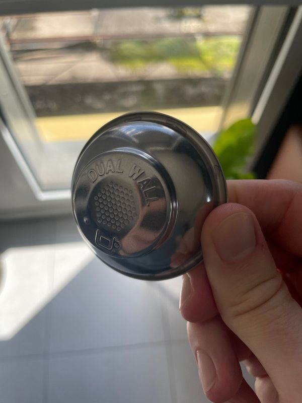 A hand holds a dual wall espresso machine basket against a bright background.