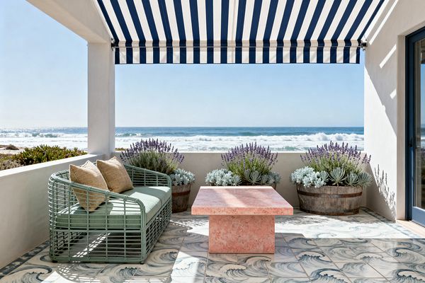 An oceanfront patio with a navy-and-white striped awning shades a seafoam green open-lattice sofa with pale green cushions and tan pillows. A square coral-pink stone coffee table sits on blue-and-white floor tiles patterned like waves. A low white stucco wall holds weathered barrel planters of lavender and silvery succulents, framing expansive sea views under bright, natural light for a breezy coastal atmosphere.