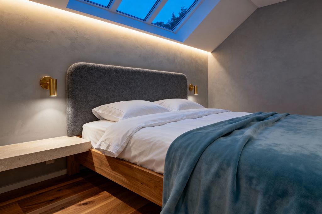 A cozy Scandinavian bedroom under a sloped ceiling with three skylight windows and concealed LED lighting. A low solid-wood platform bed with white cotton bedding and a muted teal throw sits against smooth gray plaster walls, featuring a rounded charcoal felt headboard and two brass cylinder sconces. A light limestone floating night shelf and warm wood plank flooring complete the minimal, serene space.