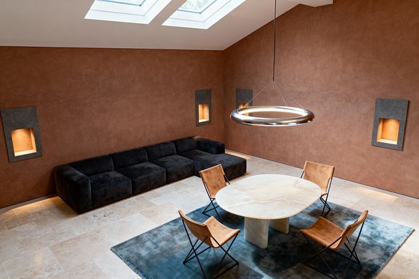A minimalist modern living room with a high, sloped ceiling and two skylights. Terracotta plaster walls with recessed, lighted niches frame a low charcoal modular sofa on pale limestone tile. At the center, an oval alabaster pedestal table sits on a smoky blue rug, surrounded by tan leather sling chairs with black metal frames and illuminated by a suspended brushed-steel circular LED ring pendant and subtle perimeter floor lighting.