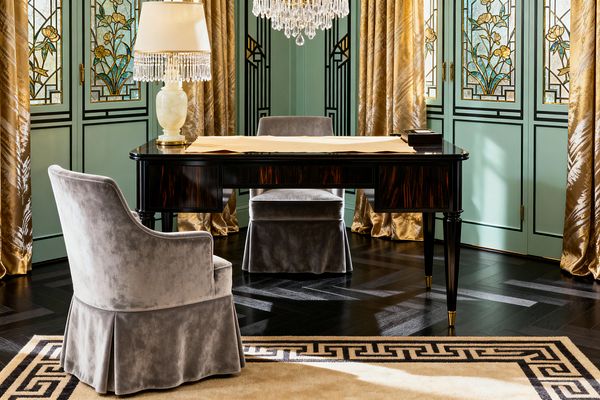 A Vintage Glam home office centers on a glossy dark wood desk with brass-capped legs and an alabaster lamp trimmed with crystal fringe, paired with two gray velvet skirted chairs. Mint green paneled doors with floral stained-glass inserts and champagne-gold drapery surround the space, while a tiered crystal chandelier hangs above a dark herringbone wood floor and a cream rug with a black Greek key border, creating a luminous, richly detailed workspace.
