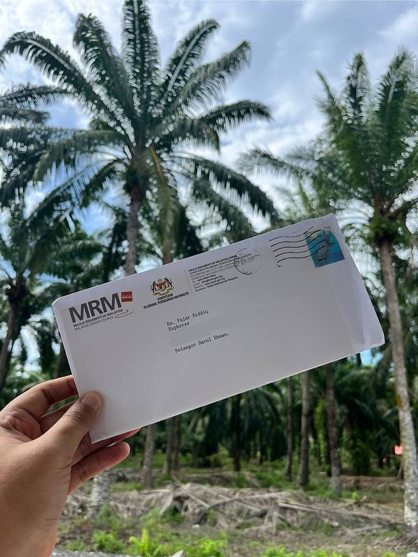 A person holds a letter in front of a palm tree landscape in Malaysia.