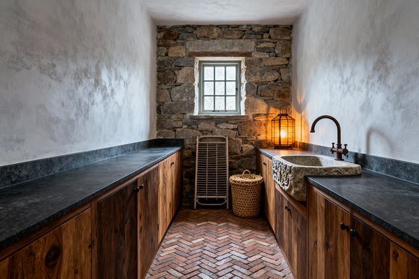 A narrow galley laundry room with knotty wood base cabinets and dark stone countertops on both sides. A carved stone farmhouse sink with a wall-mounted aged bronze faucet sits to the right, lit by a warm caged lantern. The floor is herringbone terracotta brick leading to a rough fieldstone wall with a small window. Limewashed plaster walls frame the space, with a wicker basket and wooden drying rack near the window, creating a calm, rustic look.