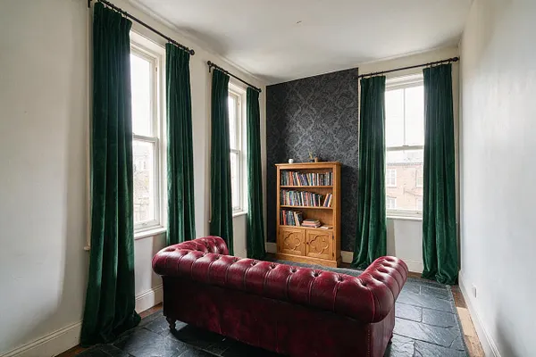 A narrow sunroom with tall windows dressed in deep green velvet curtains, a burgundy tufted leather sofa, and a small wooden bookcase against a dark patterned accent wall. The slate floor and subdued palette create a moody Gothic atmosphere with plenty of natural light.