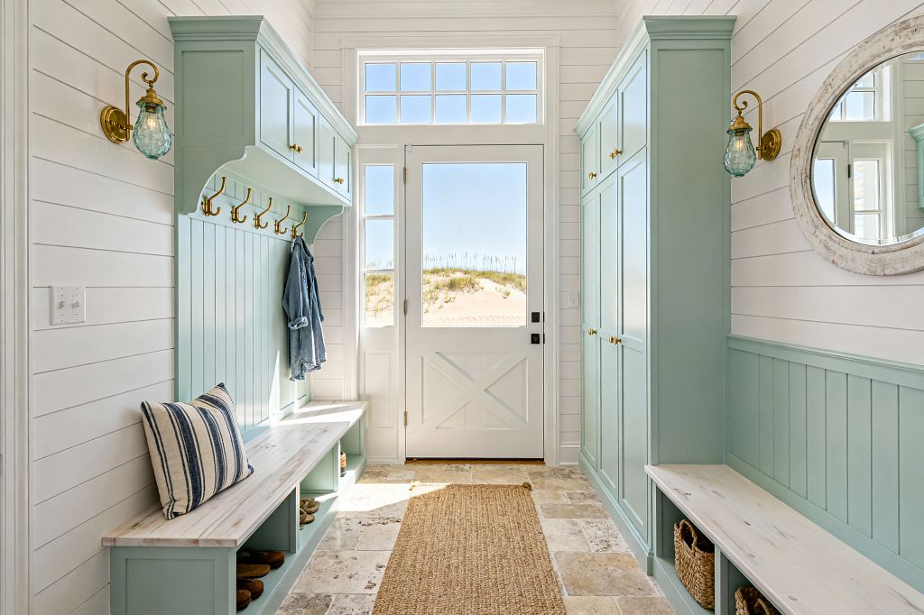 A bright coastal mudroom with white shiplap walls features seafoam green built-in lockers and benches flanking a white door with a large window and transom. Brass hooks, small brass knobs, and aqua seeded-glass sconces add warm sparkle. The benches have whitewashed wood tops and cubbies with woven baskets and shoes; a striped pillow and a denim jacket rest casually. A tumbled stone floor and jute runner lead to a view of sand dunes outside.
