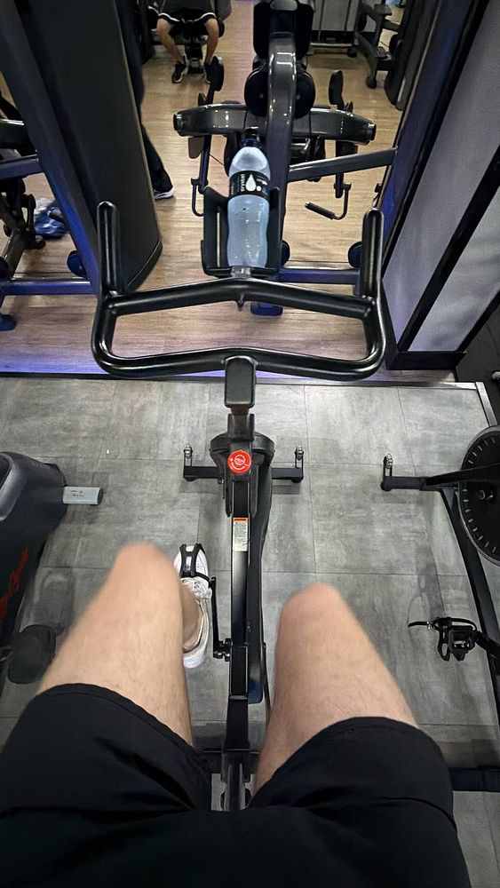 Photograph of a swimming pool with colored lane markers and a building labeled 'PISCINA' in the background under a blue sky with some clouds.