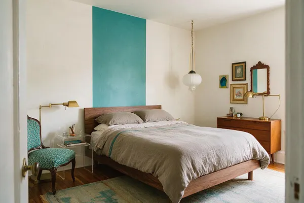 A master bedroom with a low wooden bed dressed in soft gray bedding and centered against a tall teal accent stripe on white walls. The room mixes vintage and modern pieces, including a patterned teal chair, brass task lamps, a mid-century wooden dresser, framed art, and a white pendant light.