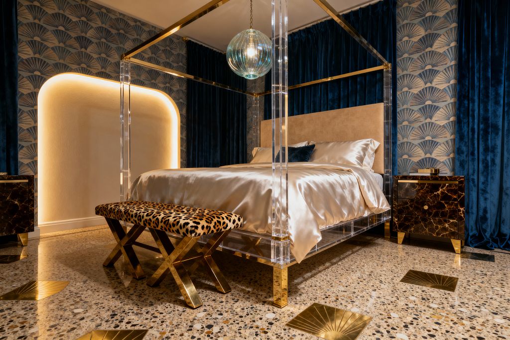 A glamorous bedroom features a clear acrylic four-poster bed with brass canopy rails, champagne satin bedding, and a beige upholstered headboard. Deep navy velvet curtains and fan-patterned Art Deco wallpaper frame a backlit arched niche, while a ribbed glass globe pendant hangs above. A leopard-print bench with brass X-legs sits at the foot, flanked by dark marble nightstands. Terrazzo flooring with brass inlays adds shine.
