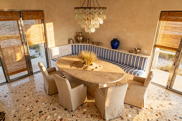 A sunlit coastal dining room features an L-shaped, whitewashed wood banquette upholstered in blue-and-white stripes. At the center sits an oval travertine table with a recessed sand planter holding beach grass, surrounded by four beige linen slipcovered armchairs. A tiered capiz shell chandelier hangs above. The floor is terrazzo embedded with seashells, while woven bamboo shades filter light through steel-framed sliding doors; silver vessels and a cobalt blue vase decorate a rear ledge.