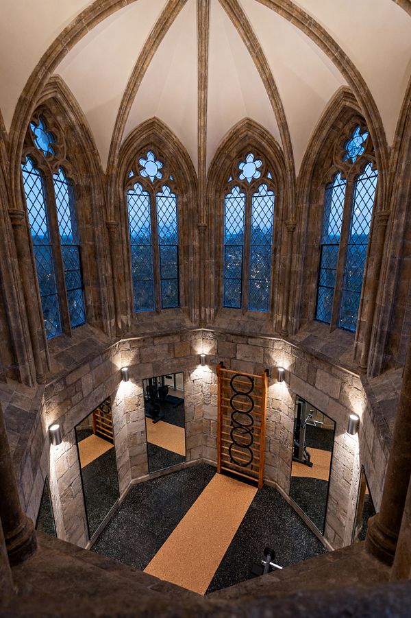 A home gym fills a small stone tower with a lofty vaulted ceiling and pointed Gothic windows glazed in leaded diamond panes with quatrefoil details. Brushed-steel sconces illuminate rough-cut masonry, full-height mirrors, an oak stall bar, and speckled black rubber flooring with a tan cork runner, while twilight views through the windows reveal trees outside, blending medieval architecture with contemporary fitness features.