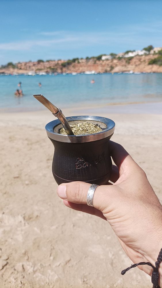 A hand holds a traditional mate cup at the beach with a scenic background.
