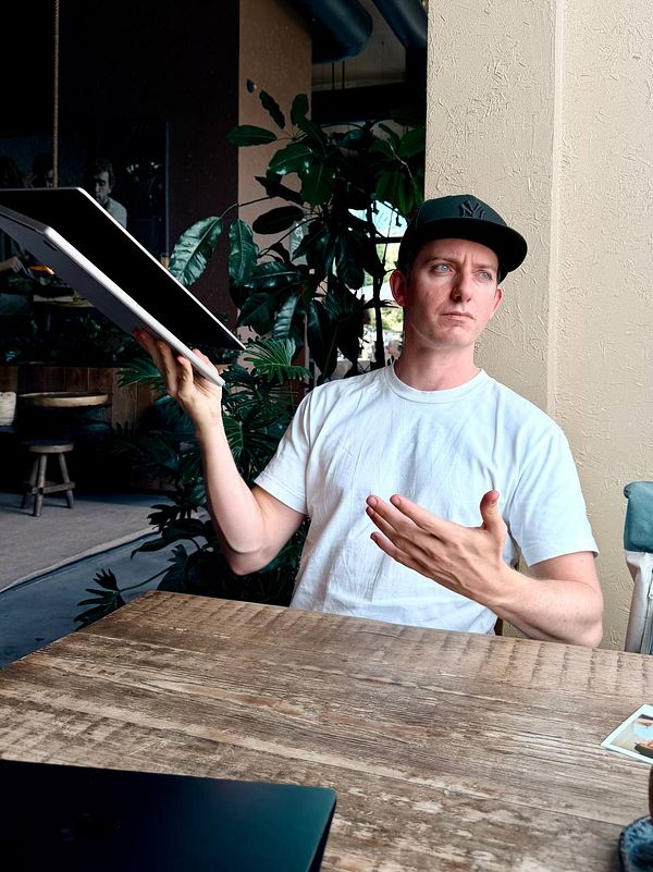 Portrait of a young man in a blue shirt captured in natural light, sitting on a wooden chair against a plain blue background.