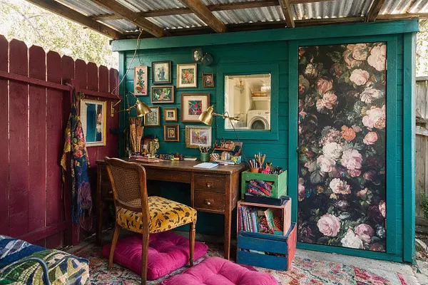 A colorful backyard studio under a corrugated metal roof with teal-painted walls and a deep burgundy fence. The space includes a wooden desk, patterned upholstered chair, framed artwork, brass task lamps, stacked storage crates, and a door covered in large floral wallpaper.