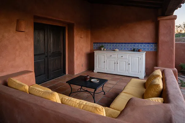 A Mediterranean-style patio with terracotta stucco walls, a dark wood double door, and a terracotta tile floor. Yellow cushioned outdoor seating surrounds a small black coffee table, while a white sideboard stands against a blue patterned tile accent strip.