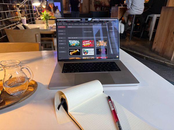 A laptop is set up on a table in a café, displaying a portfolio interface alongside a notebook and a pen.