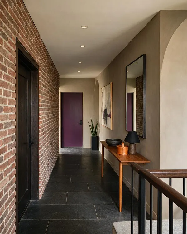A modern entryway corridor with a exposed red brick wall on the left, taupe painted walls on the right, and dark slate floor tiles. A slim wooden console table holds decorative bowls, a lamp, and a planter beneath a tall black-framed mirror, with a deep purple door at the end of the hall.